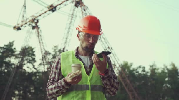 Trabalhador masculino eletricista chefe em chapéu duro tenta comer sanduíche e beber café, mas precisa trabalhar para supervisionar o processo de falar ao telefone no local de trabalho perto da linha de energia. Não há tempo para descansar — Vídeo de Stock
