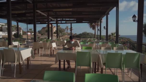 Pria bertopeng yang kesepian duduk dan menggunakan ponsel pintar sendiri di restoran kosong di Siprus di Agios Georgios. Pengunjung mengenakan topeng menunggu perintah sambil duduk di kafe kosong, jarak sosial dan karantina — Stok Video