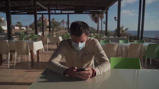 Lezárás, karantén és éttermek bezárása Cipruson. Szomorú álarcos férfi ül az üres kávézóban és telefonál az Agios Georgios kikötőben. Férfi maszkban, mobiltelefonnal a kávézóban. — Stock videók