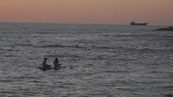 Två flickor sitter på sup board i det lugna Medelhavet vid solnedgången i staden paphos i cyprus. Silhuetter av 2 flicka paddling på paddelbräda vid solnedgången. Vattensport nära stranden vid solnedgången — Stockvideo