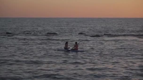 Дві жінки поділяють весло-дошку на заході сонця на спокійних водах у середземноморському морі на ципру. Дві дівчинки веслують на позашляховику. Силует вдалині. Водяний спорт, серфінг, серфінг чи весло - борт. — стокове відео
