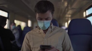 Trenle seyahat eden akıllı telefonlu maskeli bir adam. Toplu taşımada antivirüs maskesi takan beyaz bir erkek. Maskeli yolcu banliyö treninde demiryolu ile seyahat ederken akıllı telefon kullanıyor