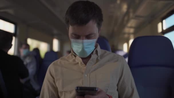 Hombre enmascarado con smartphone viajando en tren. Varón caucásico con máscaras antivirus en el transporte público. Pasajero enmascarado usa teléfono inteligente mientras viaja en tren en tren de cercanías — Vídeos de Stock