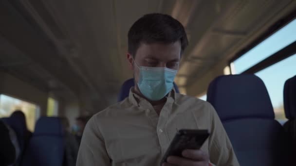 Тема новой нормы и путешествия во время пандемии коронавируса: человек в защитной маске путешествует в поезде и использует смартфон. Счастливого пути. Социальное расстояние в общественном транспорте — стоковое видео