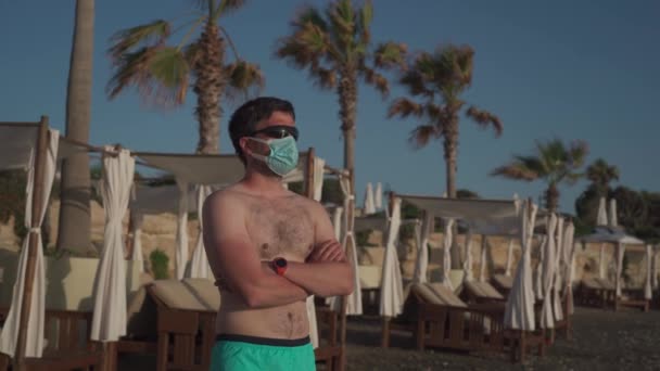 医療用マスクの男は、コロナウイルスの隔離とロックダウンの間、キプロス、パフォス市のサンラウンジャーとヤシの木の近くの空のビーチに一人で立っています。COVID-19パンデミック発生後の休暇中の新しい正常 — ストック動画