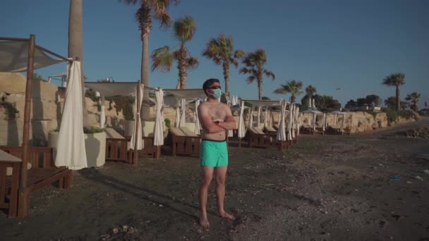 Riposare l'uomo in una maschera protettiva su una spiaggia sporca da solo sullo sfondo di palme e lettini sulla costa mediterranea a Cipro. maschio avendo vacanza estiva durante covid blocco — Video Stock