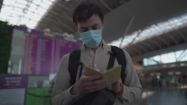 Man resenär i medicinsk mask kontrollera pass på flygplatsen mot bakgrund styrelse med schema för avgångar och ankomster. Man med ryggsäck gärna resa igen, öppna gränser efter coronavirus — Stockvideo