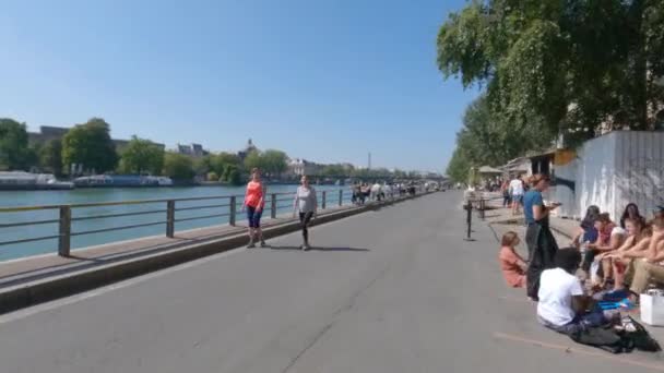 Набережная на краю канала с пешеходами. Туристы и парижане ходят пешком, ездят на велосипедах и едят в кафе рядом с рекой Сена в жаркий летний солнечный день. Передвижная камера. Париж, Франция 2 сентября 2021 года — стоковое видео