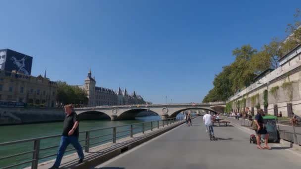 Embankment at edge of canal with pedestrians. Tourists and Parisians walk, ride bicycles and eat in cafe next to river Seine on hot summer sunny day. Moving camera. Paris, France September 2, 2021 — Stock Video