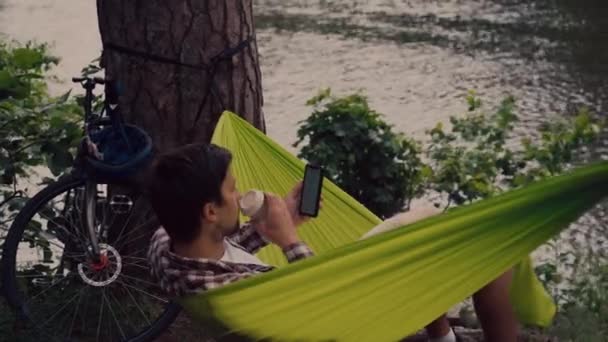 Freelancer al aire libre descansando, pausa para tomar un café en el parque después de andar en bicicleta. Hombre caucásico ciclista usando el teléfono y beber café llevar en hamaca cerca del lago. Disfrutando de las vacaciones en la naturaleza — Vídeos de Stock