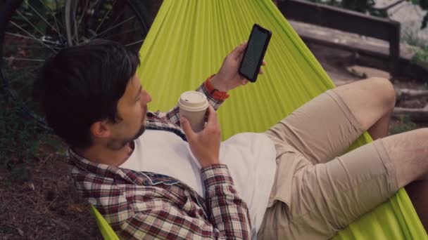 O homem navega na Internet no smartphone enquanto bebe café com ele enquanto está sentado na rede verde com vista para o lago em um parque florestal tendo chegado de bicicleta. Ciclista em rede no parque de campismo por rio — Vídeo de Stock