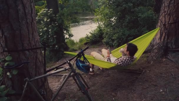 A young man came to the park on a bicycle and is resting in a hammock overlooking the lake while drinking coffee to go and surfing the Internet on a smartphone. Biking and camping concept — Stock Video