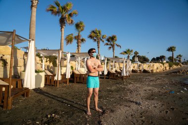 Tıbbi maskeli adam, Coronavirus karantinası ve tecrit sırasında Kıbrıs, Paphos şehrinde güneş pansiyonları ve palmiye ağaçları yakınlarındaki boş sahilde tek başına duruyor. COVID-19 salgınından sonra tatilde yeni normal.