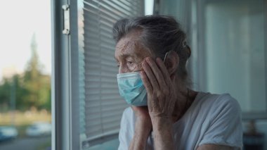 90 yaşında koruyucu maske takmış üzgün bir yaşlı kadın huzurevinin açık penceresinden dışarı bakıyor. Yüzü maskeli yalnız yaşlı bir kadın endişeyle pencerenin kenarında duruyor, elleriyle yüzünü kaldırıyor.