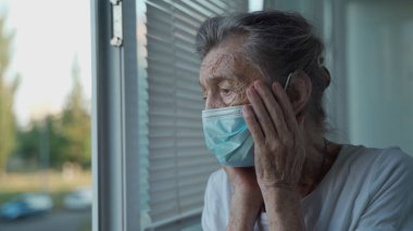Koruyucu tıbbi maske takmış endişeli yaşlı bir kadın üzgün bir şekilde pencereden dışarı bakar. Kafası bir huzurevinde elleri kelepçelidir. Covid-19 salgını sırasında evde depresyondaki bir kadın vardı.
