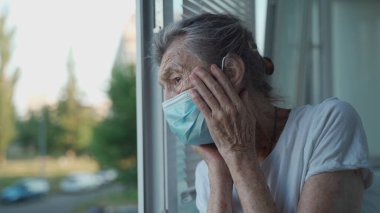 90 yaşında koruyucu maske takmış üzgün bir yaşlı kadın huzurevinin açık penceresinden dışarı bakıyor. Yüzü maskeli yalnız yaşlı bir kadın endişeyle pencerenin kenarında duruyor, elleriyle yüzünü kaldırıyor.