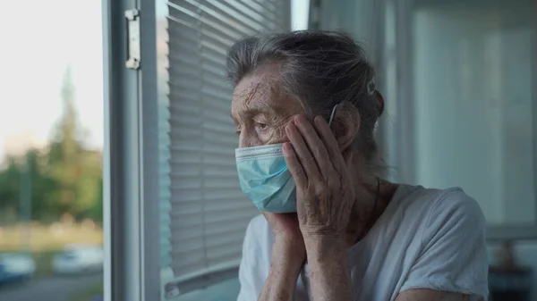 Mujer mayor triste de 90 años con máscara médica protectora mirando por una ventana abierta en un asilo de ancianos. Solitaria anciana enmascarada en la cara se para ansiosamente junto a la ventana, levanta las manos para enfrentarse —  Fotos de Stock