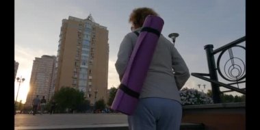 Formda yürüyen yaşlı bir kadın. Açık hava yoga dersine giden mutlu olgun bir bayan. Aktif yaşam tarzı. Salgından sonra normal hayatın yeniden başlaması. Egzersiz paspasıyla yürüyen yaşlı bir kadın.