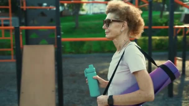 Mujer mayor activa con tapete de fitness y botella de agua en las manos caminando en el gimnasio al aire libre de entrenamiento. Feliz señora madura paseando por el campo de deportes yendo a clases de yoga al aire libre. Concepto de estilo activo — Vídeos de Stock