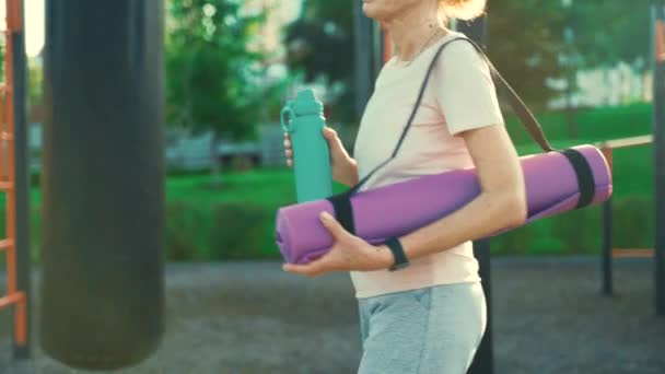 Actieve seniorenvrouw met fitnessmat en waterfles in handen lopen op workout outdoor fitnessruimte. Gelukkig volwassen dame wandelen in sportveld gaan naar buiten yoga klasse. Actieve levensstijl — Stockvideo