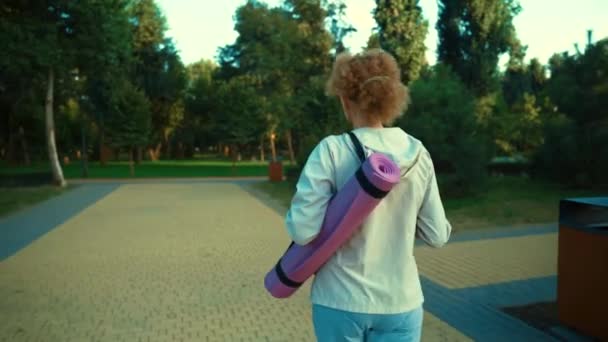 Mujer mayor con alfombra de fitness caminando. Feliz señora madura que va a clases de yoga al aire libre. Estilo de vida activo. Reinicio de la vida normal después de una pandemia. Mujer mayor caminando con alfombra de ejercicio en el entrenamiento — Vídeo de stock