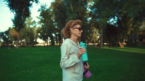 Mujer madura yendo a clases de yoga al aire libre. Senior activo disfrutando de un estilo de vida saludable. Mujer mayor sosteniendo alfombra de ejercicio mientras camina en la sesión de entrenamiento. Mujer anciana positiva con equipo deportivo — Vídeos de Stock