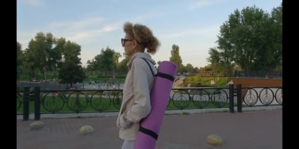 Mujer madura yendo a clases de yoga al aire libre. Senior activo disfrutando de un estilo de vida saludable. Mujer mayor sosteniendo alfombra de ejercicio mientras camina en la sesión de entrenamiento. Mujer anciana positiva con equipo deportivo — Vídeos de Stock