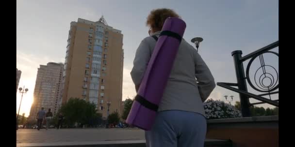 Senior vrouw met fitness mat lopen. Gelukkige volwassen dame gaat naar buiten yogales. Actieve levensstijl. Herstart normaal leven na een pandemie. Oudere vrouw wandelen met een trainingsmat op de training — Stockvideo