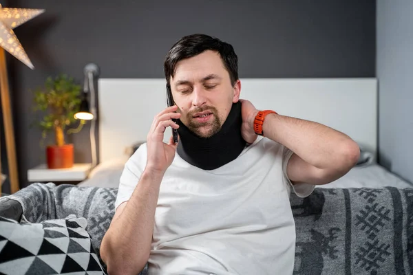 Disabled man with neck brace talking on phone with doctor, experiencing pain while sitting on couch in the living room at home. Injured young man using cervical brace makes calls using mobile phone.