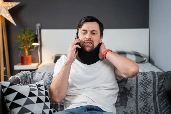 Man in foam cervical collar is calling on phone to the insurance company for medical assistance after accident, feeling neck pain, injury and suffering at home. A male with neck injury calls doctor.