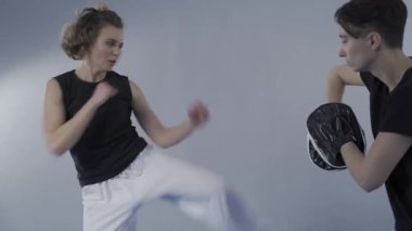 Çıplak ayaklı bir kadının boks patisi tutan tekvando koçuyla tekmeleme alıştırması yaptığı yakın plan. Spor salonunda kişisel koçuyla boks kalkanını tekmeleyen konsantre kadın. Dövüşçüler birlikte çalışır..