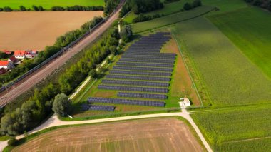 Luftaufnahme mit Drohne uber Bayern, Deutschland. Bahngleise fuehren durch Felder vorbei bir einer Solaranlage. Solarpaneele glaenzen in Sonne und zeigen nachhaltigen Verkehr und erneuerbare Energie