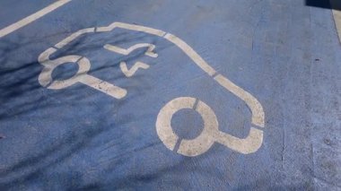 Mavi asfalta elektrik yükleme sembolü çizilmiş. Otoparkı şarj eden bir EV var. Sürdürülebilir hareketlilik, temiz enerji ve modern elektrik taşımacılığı için kentsel altyapı kavramı