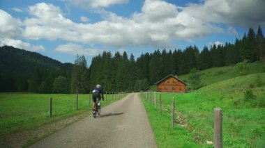 Çakıl taşı bisikletli bir bisikletçinin kaldırımlı yolda, manzaralı Bavyera ya da Avusturya Alpleri 'nden geçişi. Yol yeşil çayırlardan geçerek geleneksel ahşap kulübeye ve mavi gökyüzünün altındaki sık ormana gidiyor.