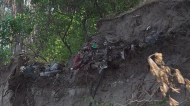 Çökmüş bir nehir kıyısına gömülü çöp ekolojik ihmali simgeler. Gömülü atıkların erozyon, çevre kirliliği ve su kaynaklarından dolayı nasıl yüzeye çıktıklarının rahatsız edici bir görüntüsü