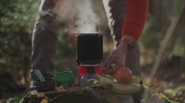 Entegre gaz pişirme sistemi sobası ve tencere kombinasyonunu sonbahar ormanlarında ayarlayan yürüyüşçü, verimlilik, yakıt tasarrufu ve açık hava macerası için kompakt teçhizatı vurguluyor. Bir kütükte muz ve bir portakal.
