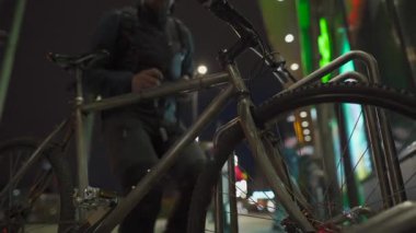 Şehir geceleri vızıldarken, bir bisikletçi bisikletini bir alışveriş merkezinin yakınındaki iyi aydınlatılmış bir alanda hırsızlık önleyici bir kilitle kilitliyor. Şehir güvenliği, güvenli park yeri ve modern metropolde hırsızlık önleme.