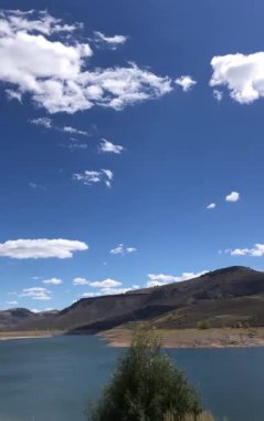 Dikey POV sürücüsü Colorado 'daki Gunnison Nehri' ni gösteriyor. Bir rezervuarın sakin mavi suyu kuru yuvarlanan tepelerle çevrilidir. Parlak mavi bir gökyüzünün kabarık beyaz bulutları vardır..