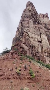 Dik durağan alçak açı videosu Utah 'taki Zion Ulusal Parkı' nda bulutlu bir gökyüzüne doğru yükselen büyük bir kumtaşı uçurumu gösteriyor. Kaya yüzeyinin alt yamaçlarında büyüyen yeşil çalılar var..