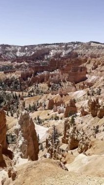 Dikey görüntüler Bryce Canyon Ulusal Parkı amfitiyatrosunu gözler önüne seriyor. Turuncu ve beyaz kabadayılar, mavi gökyüzünün altındaki toprak bir patikada çam ağaçları ve yürüyüşçülerle manzarayı işaretliyor..