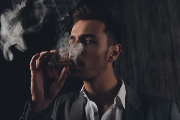 Uomo sfacciato in giacca e cravatta sullo sfondo grigio fumando un sigaro — Foto Stock