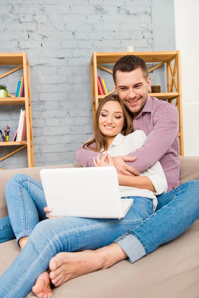 Handsome man embracing his girlfriend sitting on sofa with lapto — Stock Photo, Image