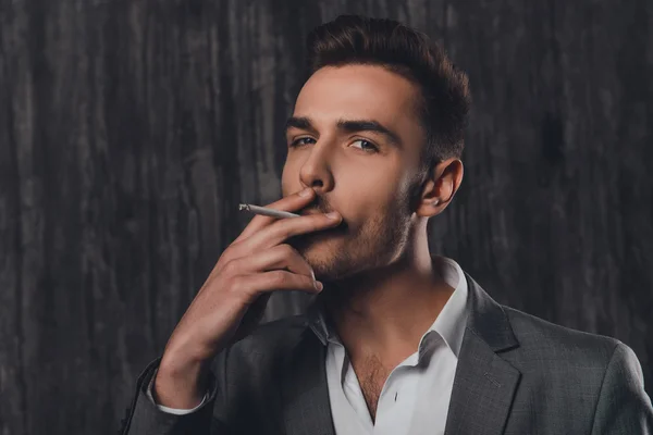 Handome brutal man in suit on the grey background smoking a ciga — Stock Photo, Image
