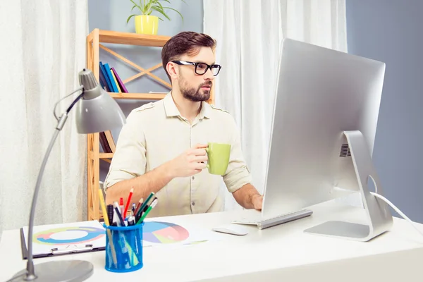 Ο άνθρωπος στο glassses πίνετε καφέ ενώ δουλεύει σε γραφείο με υπολογιστή — Φωτογραφία Αρχείου