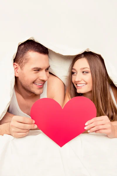 Ευτυχισμένο ζευγάρι που βρίσκεται κάτω από την κουβέρτα και κρατώντας προσφορά χαρτί καρδιά — Φωτογραφία Αρχείου
