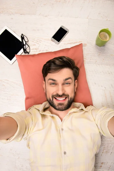 Hombre guapo acostado en el suelo en la almohada y haciendo selfie — Foto de Stock