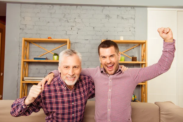 Dos hombres viendo televisión en casa con las manos levantadas — Foto de Stock