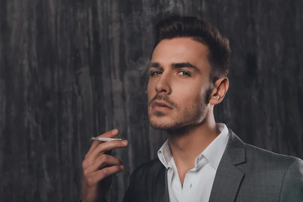 Handome uomo d'affari in giacca e cravatta fumare una sigaretta su sfondo grigio — Foto Stock