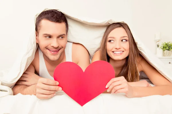 Dos amantes acostados debajo de la manta y sosteniendo el corazón de papel rojo —  Fotos de Stock