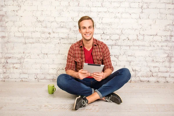 Χαρούμενος νεαρός άνδρας κάθεται στο πάτωμα με το tablet και το φλιτζάνι του καφέ — Φωτογραφία Αρχείου